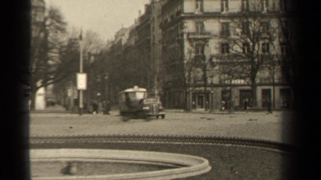 1947 Paris home-movie frame showing a passing vehicle near a formal park edge, apartment buildings, bare trees, and a circular bed.