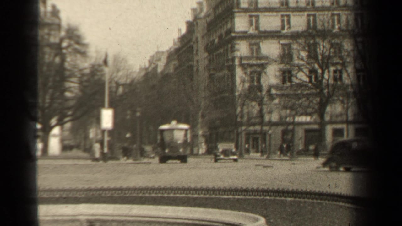 Street traffic passes in front of multi-story buildings, with bare trees and pedestrians along the sidewalk.