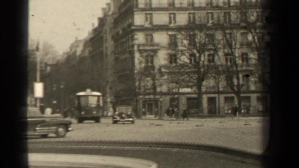 1947 Paris street view with a bus-like vehicle, cars, distant pedestrians, bare trees, and apartment facades framed by 8mm gate shadows.