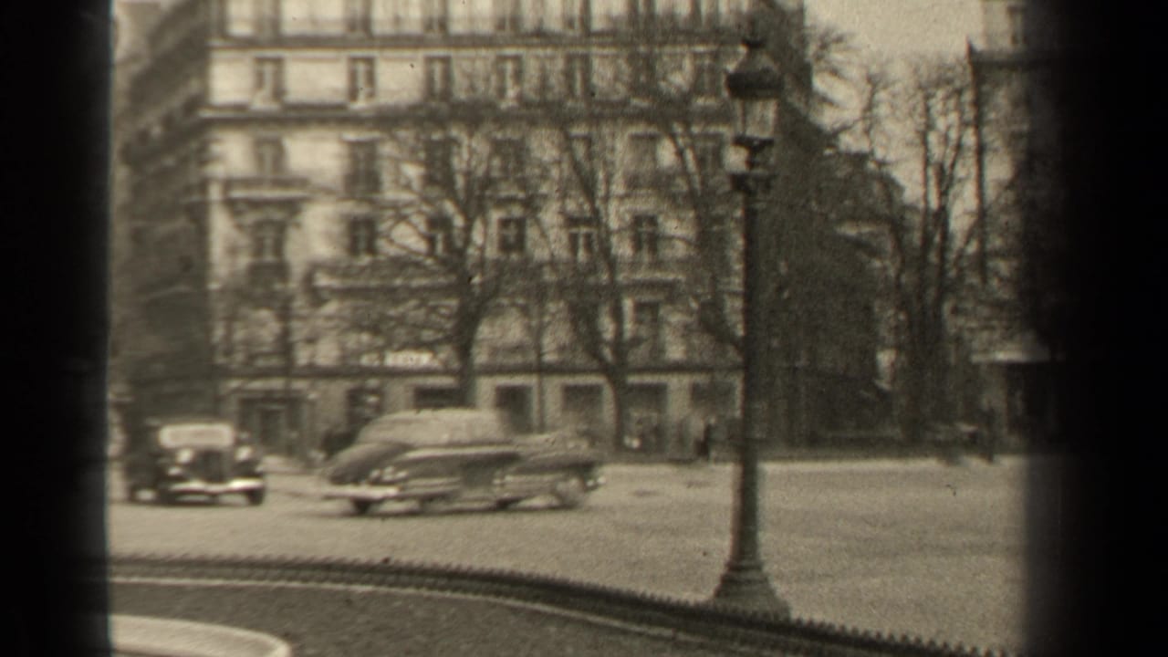 A 1947 Paris street frame with a lamppost, passing sedans, bare trees, pedestrians, and apartment buildings in grainy 8mm monochrome.
