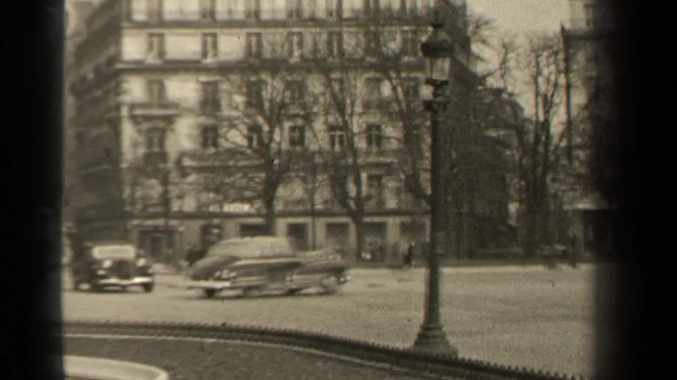 A 1947 Paris street frame with a lamppost, passing sedans, bare trees, pedestrians, and apartment buildings in grainy 8mm monochrome.