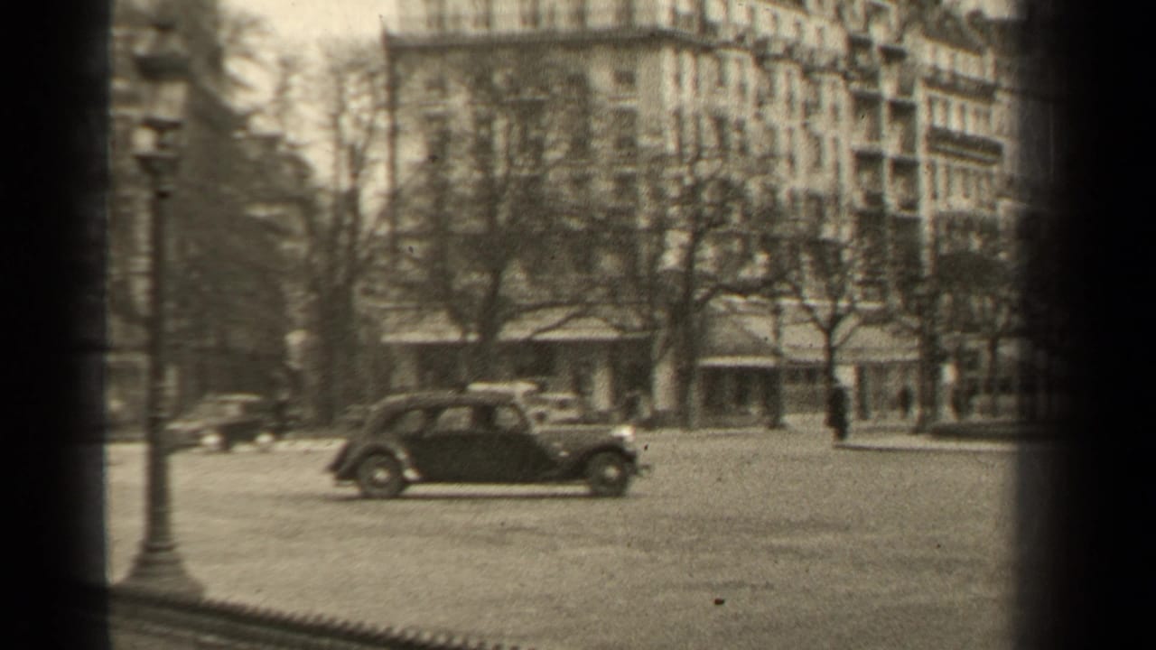 A dark car crosses an open city square in front of buildings and winter-bare trees.