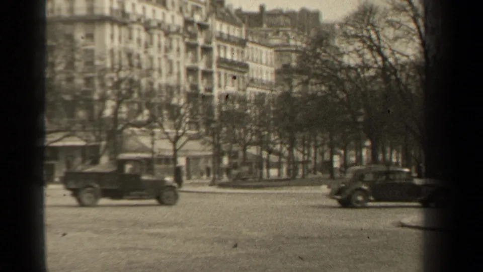 Paris street traffic in 1947, with two classic cars, leafless trees, storefronts, and residential buildings seen from a handheld home-movie viewpoint.