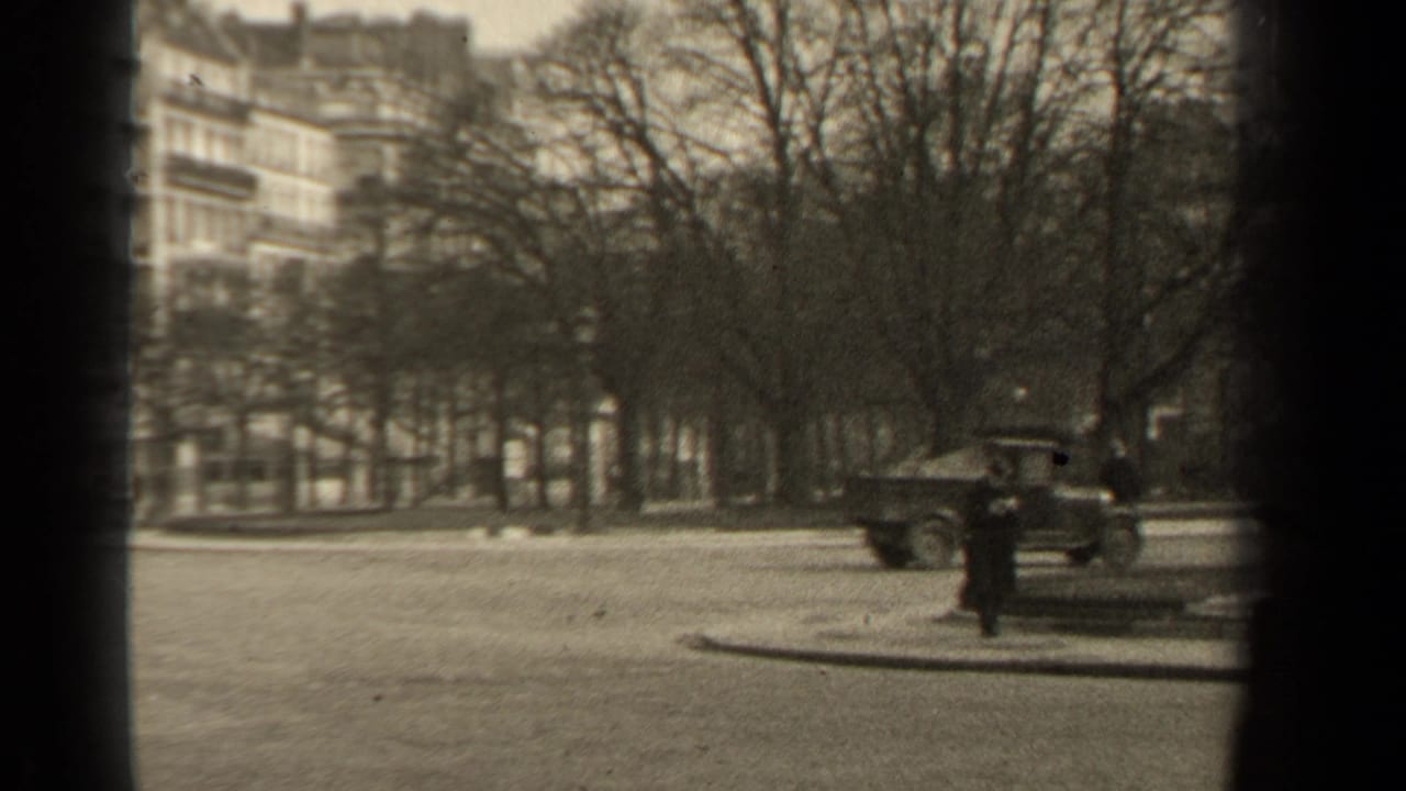 A Paris street corner with a classic car, a distant pedestrian, bare trees, apartment buildings, and a lamppost in grainy 8mm footage.