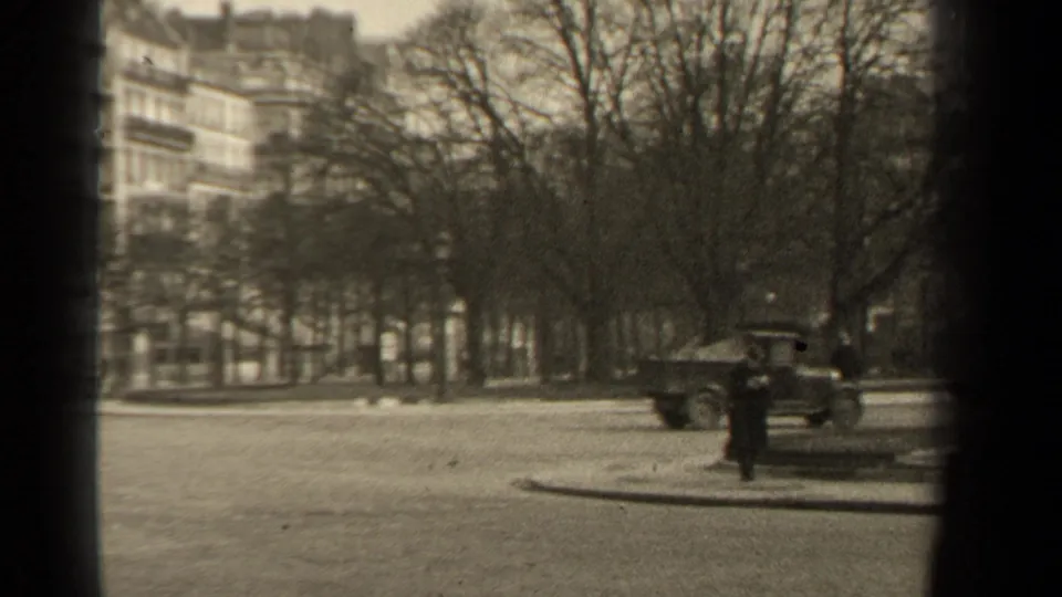 A Paris street corner with a classic car, a distant pedestrian, bare trees, apartment buildings, and a lamppost in grainy 8mm footage.