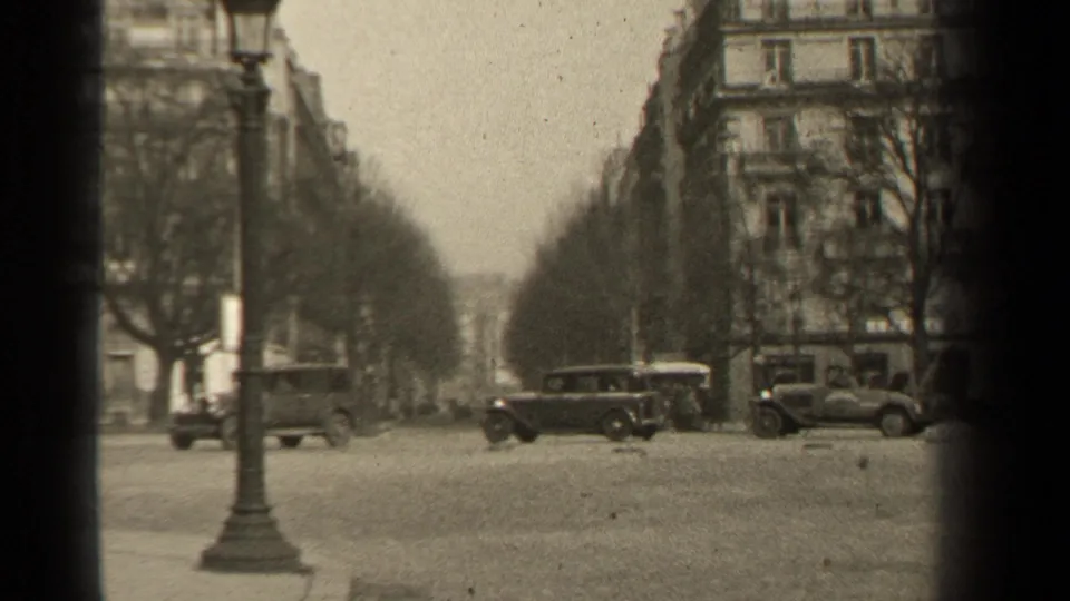 1947 Paris boulevard view with several automobiles crossing or parked near a lamppost and stone apartment blocks.