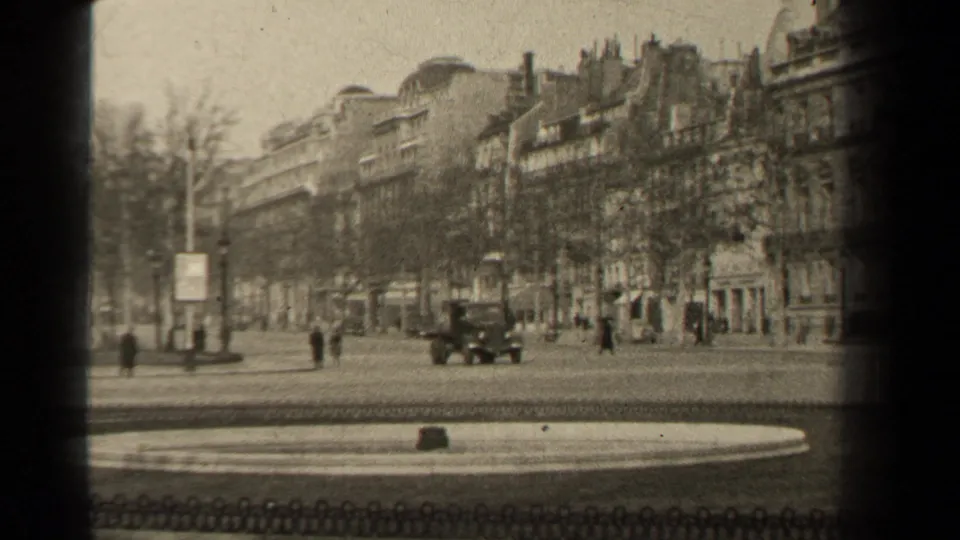 Period automobile moving through a Paris boulevard scene, with pedestrians, bare trees, and multi-story stone buildings in the background.