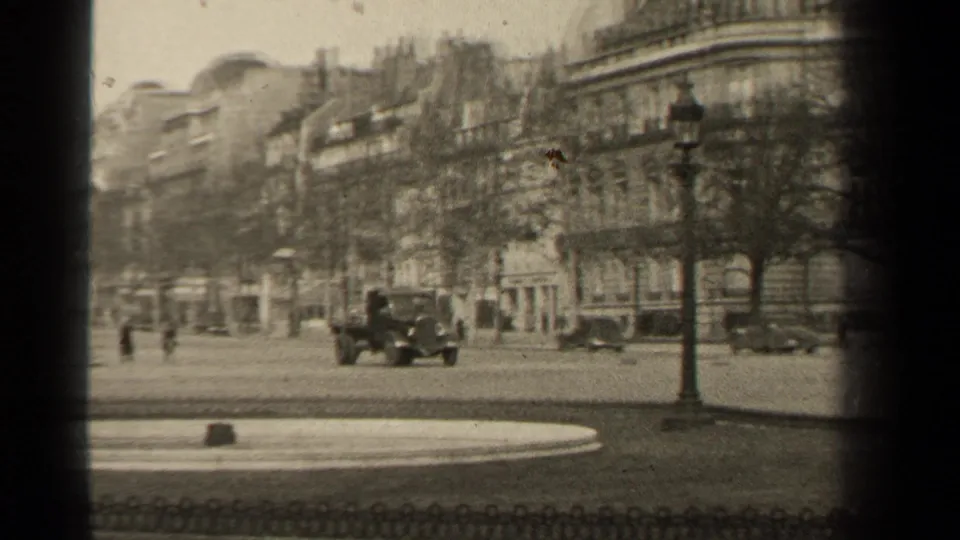 Paris street scene with multiple period vehicles, a lamppost, pedestrians, and rows of stone buildings recorded on black-and-white 8mm film.