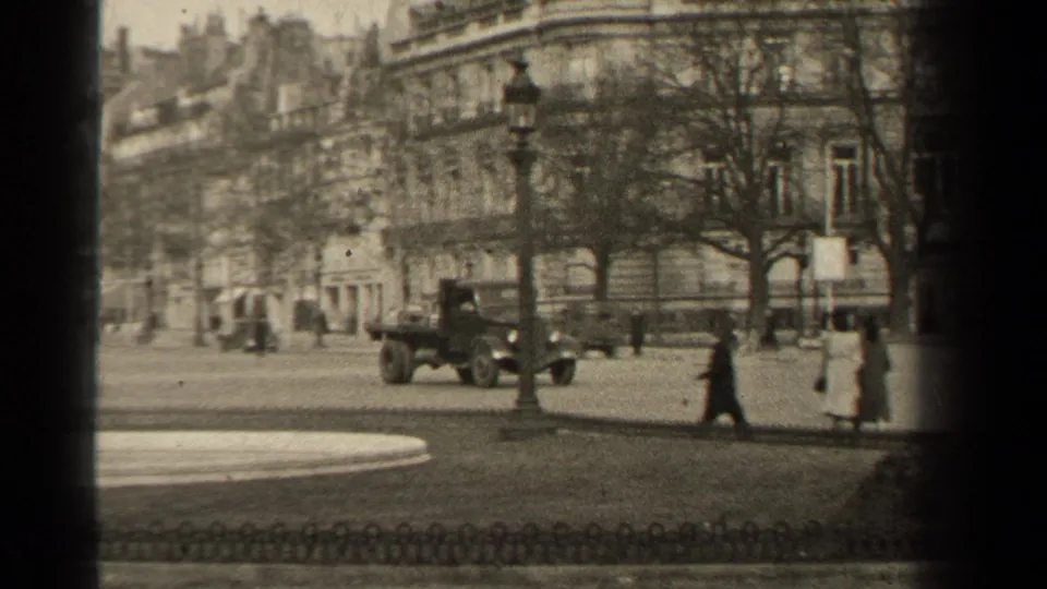 A 1947 Paris street view with pedestrians in long coats, a passing vehicle, a lamppost, bare trees, and stone facades in soft 8mm focus.