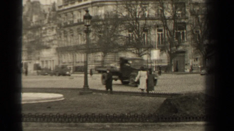 1947 Paris street view with pedestrians in coats, a boxy truck near a lamppost, classic cars, bare trees, and stone-front buildings.