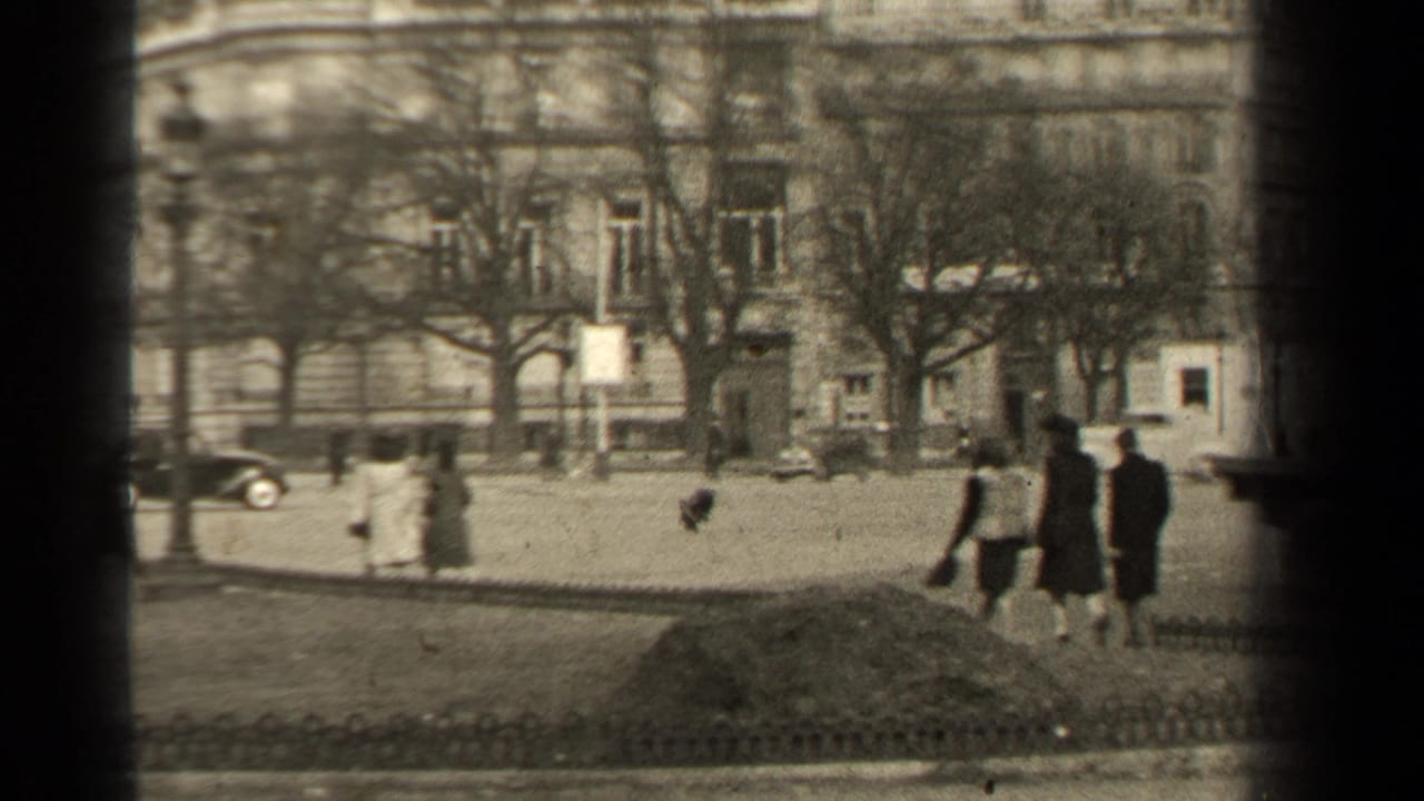 Pedestrians walk beside a broad street with classic cars, trees, and a large stone building.