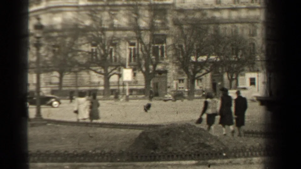 1947 Paris boulevard view with pedestrians crossing or walking near classic cars, bare trees, and a large stone building facade.
