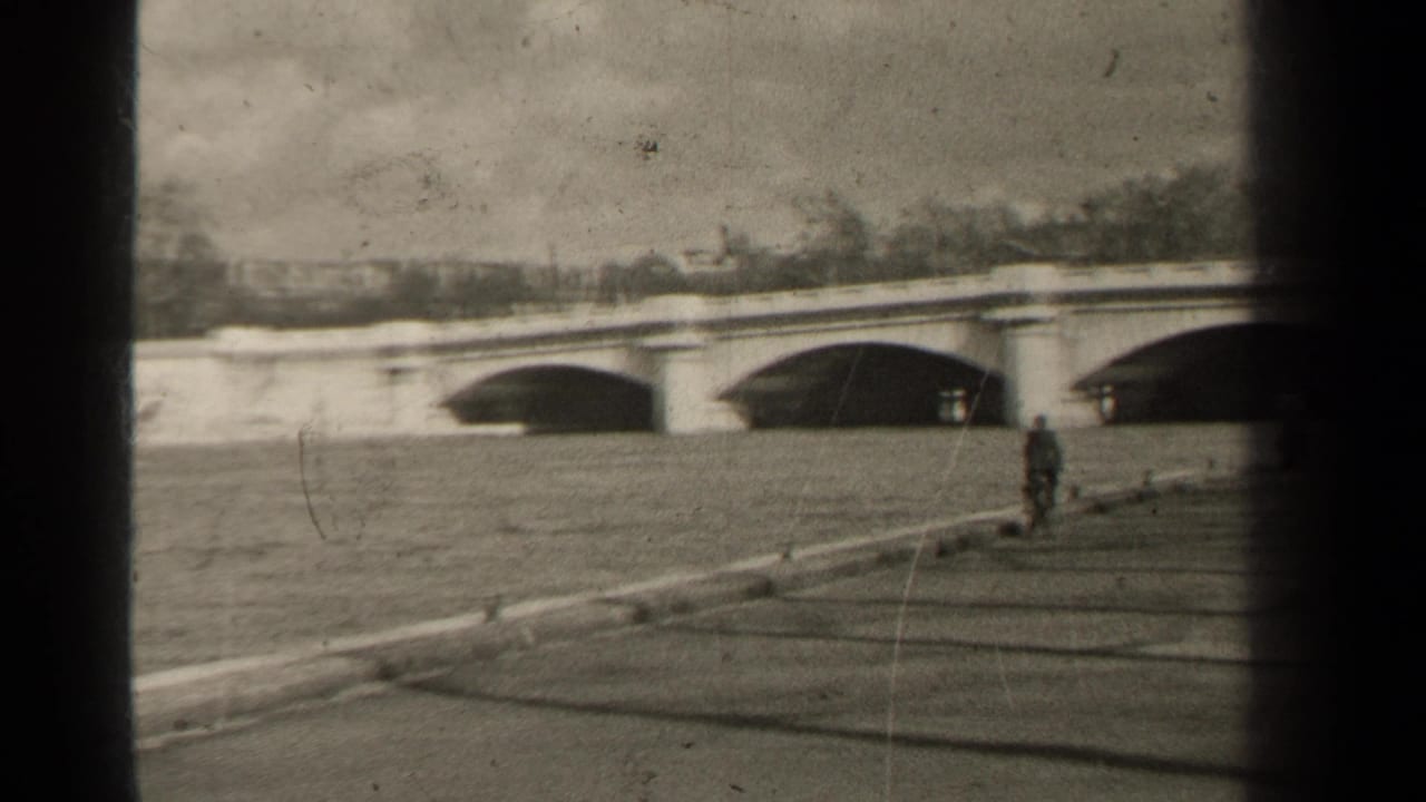 1947 Paris riverside view with an arched stone bridge, a person on the embankment, low wall, water, and distant winter trees.