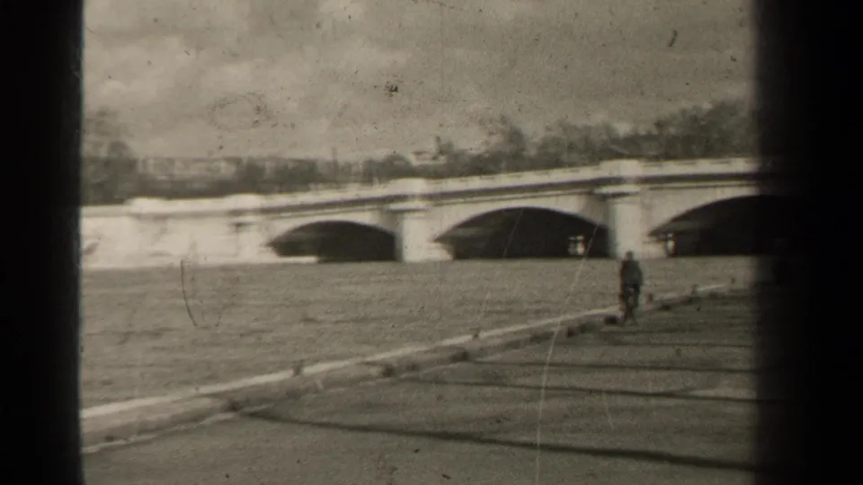 1947 Paris riverside view with an arched stone bridge, a person on the embankment, low wall, water, and distant winter trees.