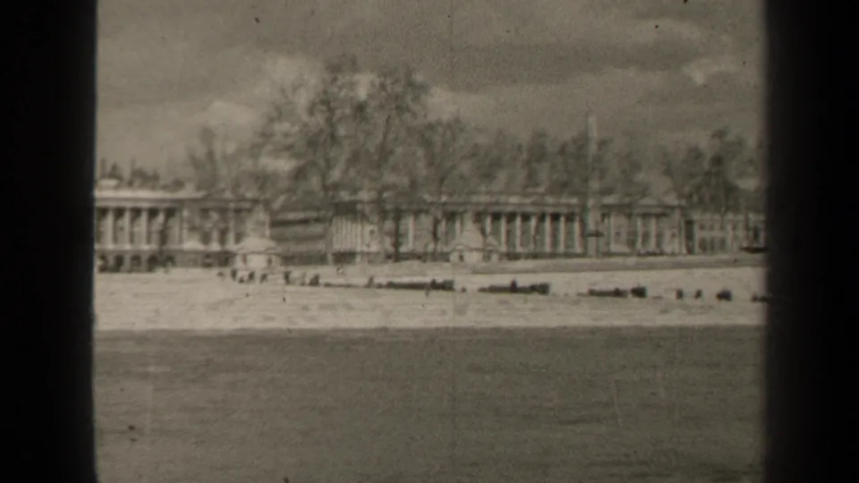 1947 Paris home-movie frame showing a long columned building beyond the Seine, with bare trees and soft 8mm contrast.