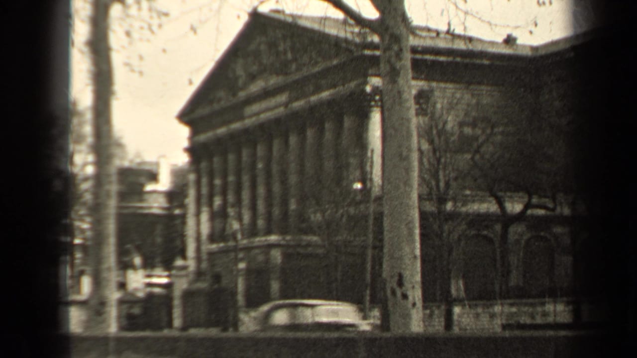 Columned building with bare trees and a light-colored car passing on the road.