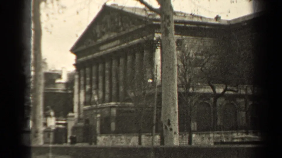 1947 Paris home-movie frame showing a grand classical building facade with columns, pediment, street edge, and bare trees.