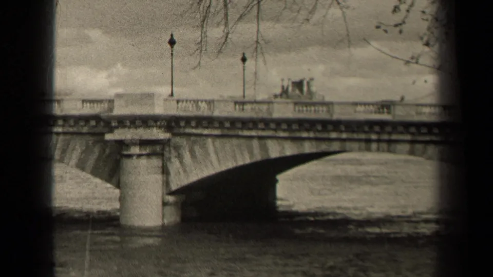 1947 Paris 8mm frame showing a stone bridge crossing a calm waterway, with lampposts, bare tree branches, and distant traffic.