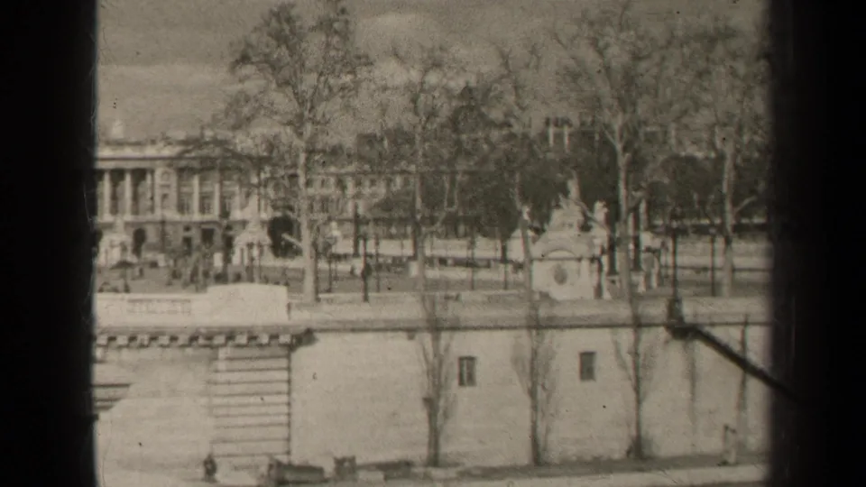 Paris home-movie frame showing a classical colonnaded building behind bare trees, with a stone embankment in the foreground.