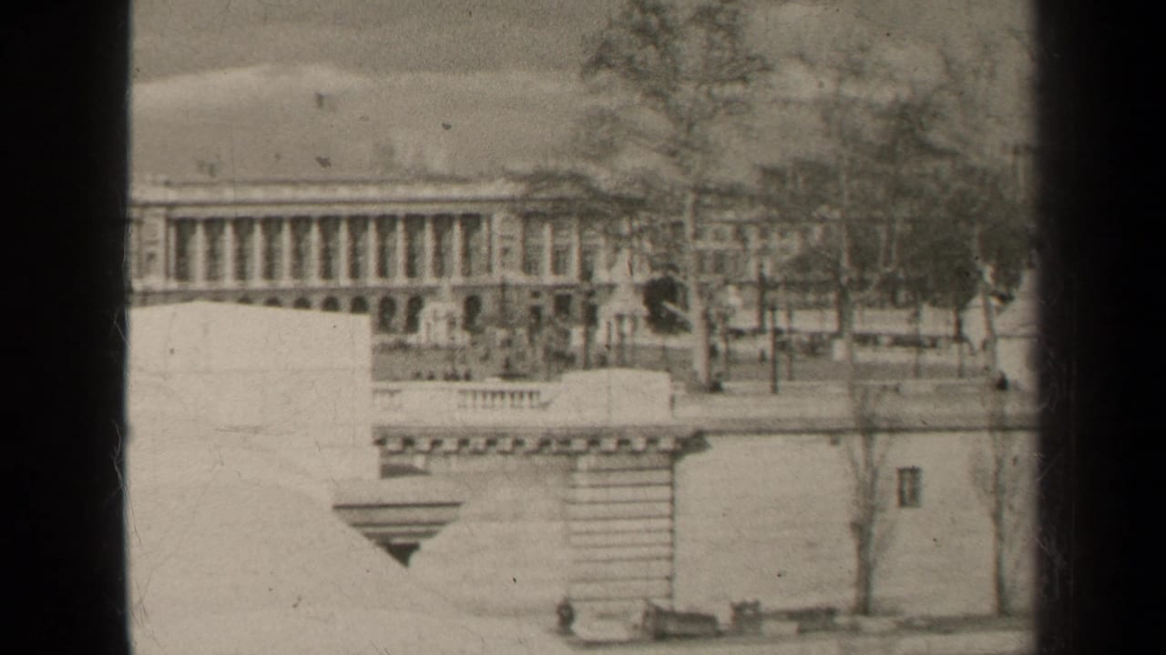 1947 Paris home-movie frame showing a formal columned building beyond stone walls, trees, and a mostly empty street.