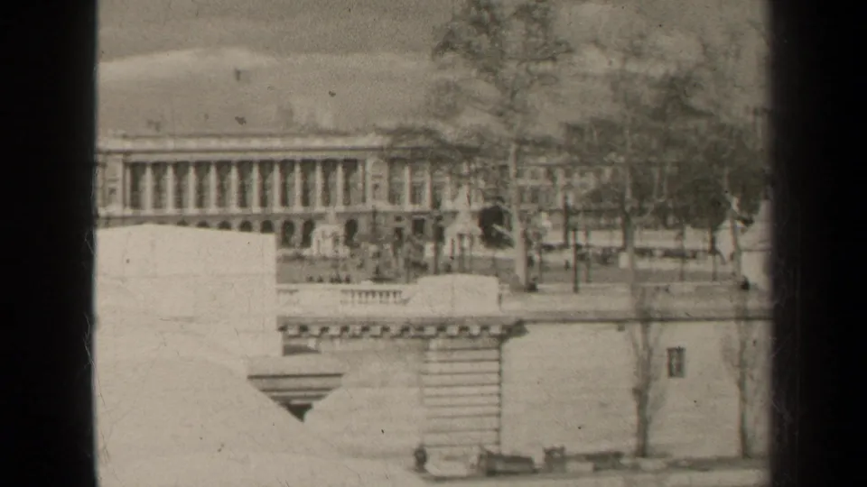 1947 Paris home-movie frame showing a formal columned building beyond stone walls, trees, and a mostly empty street.