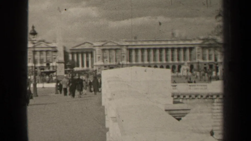 1947 Paris frame showing pedestrians crossing a public square or street near an obelisk, lamppost, and formal columned building.