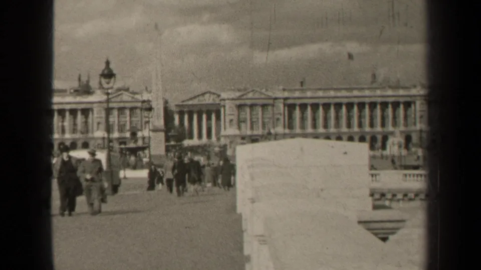 1947 Paris street scene with pedestrians in period dress near a lamppost, obelisk, stone wall, and large columned building.