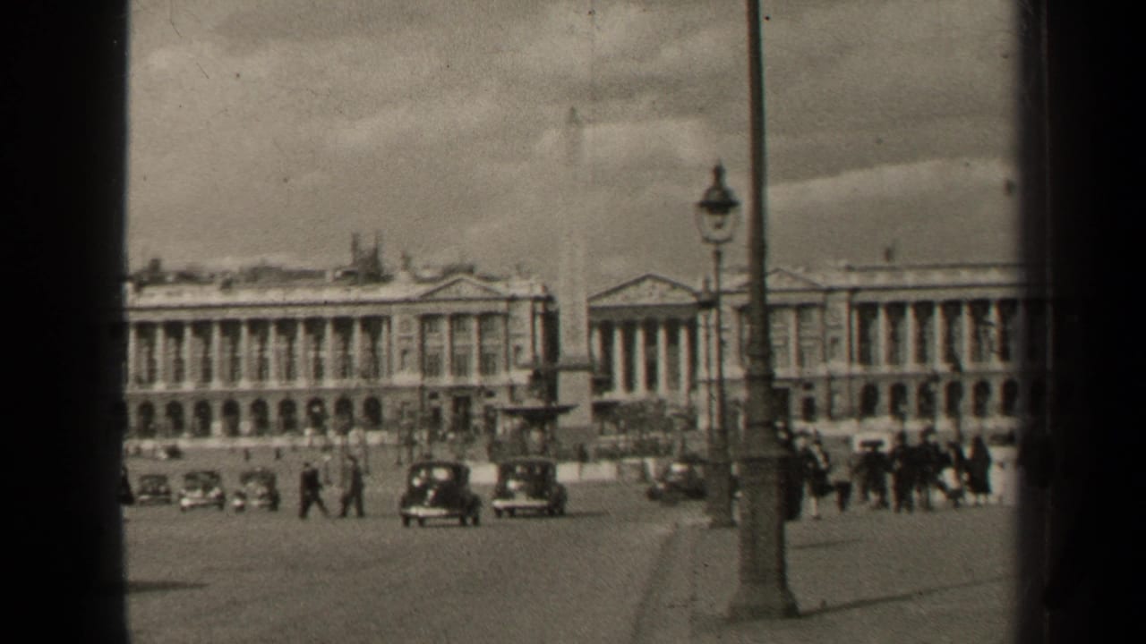 Classic cars and pedestrians cross a Paris square before long columned buildings.