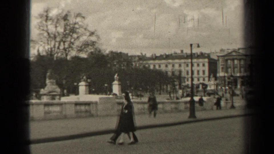 1947 Paris frame showing a couple walking beside the open square, with railings, trees, and large buildings behind them.