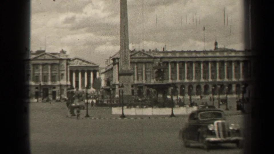 1947 Paris square scene with a foreground vintage car, obelisk, lampposts, pedestrians, and classical architecture in soft 8mm focus.