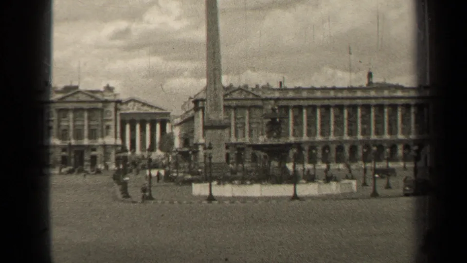 1947 8mm view of a Paris square with a tall obelisk, formal buildings, scattered pedestrians, and visible film-frame edges.