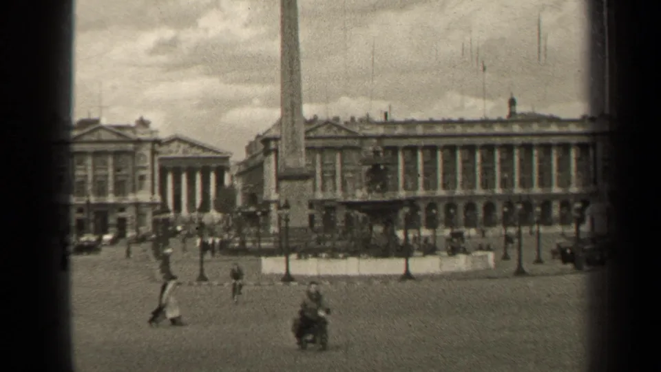 Grainy 8mm 1947 Paris frame with pedestrians and a small wheeled figure moving across a broad square before an obelisk and formal building facades.