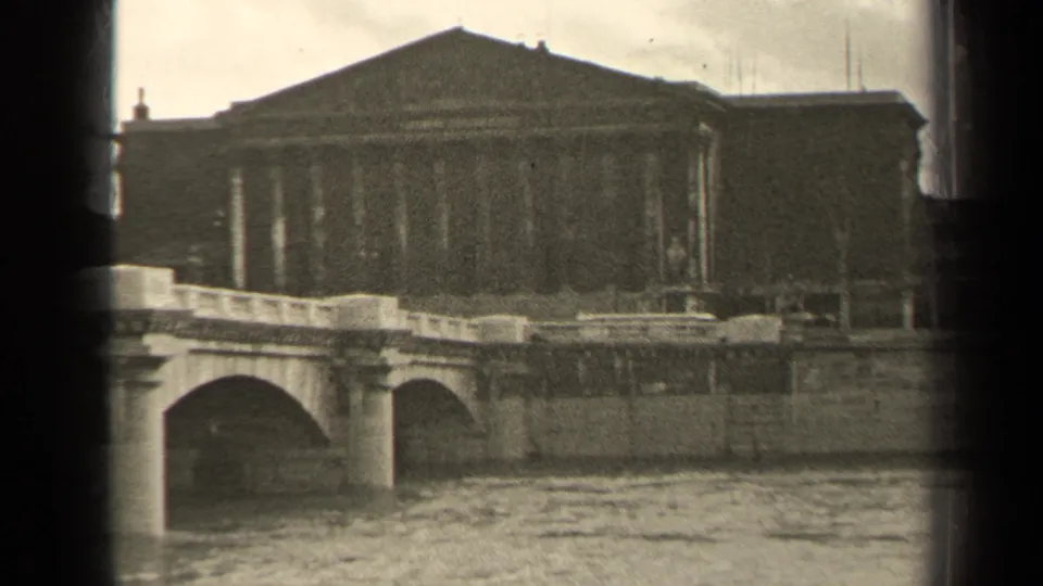 1947 Paris frame showing a river bridge and monumental columned architecture through grainy 8mm texture.