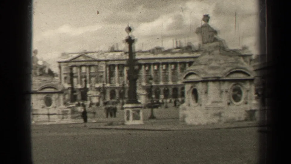 1947 Paris frame showing a statue pedestal, tall lamppost, classical building frontage, small pedestrians, and a broad street.