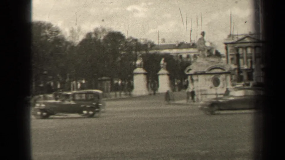 1947 Paris 8mm frame with two cars crossing a wide street, stone monuments, trees, pedestrians, and part of an ornate building.