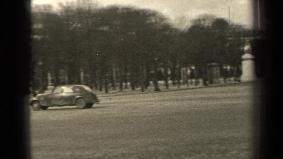 1947 Paris frame showing a rounded classic car on a wide roadway with trees, stone monuments, pedestrians, and soft 8mm grain.