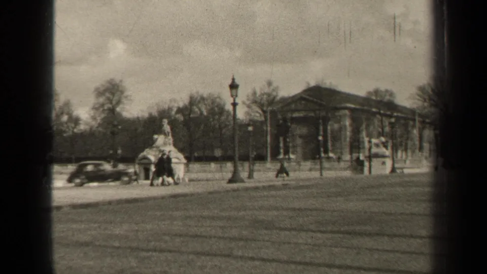 1947 Paris home-movie frame with a classic car, distant pedestrians, a small monument, lampposts, and a columned building.