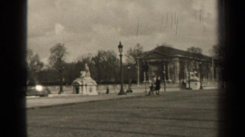 1947 Paris home-movie frame showing a soft black-and-white boulevard view with pedestrians, cars, monument, trees, and columned architecture.