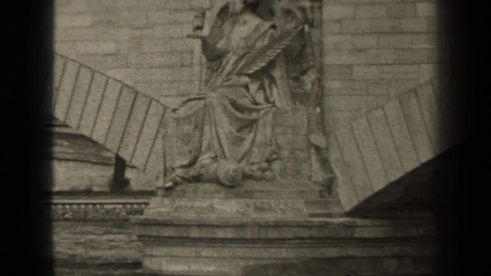1947 Paris home-movie frame of a draped seated sculpture on a plinth, with arched masonry behind it and dark film edges visible.