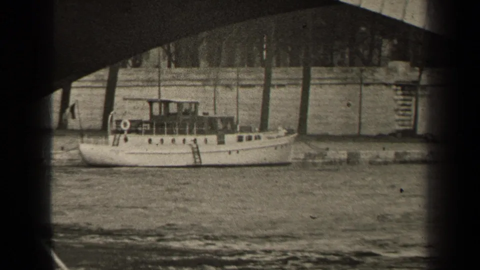 A grainy 1947 Paris home-movie view of a white boat beside the stone embankment, framed by the underside of a bridge.