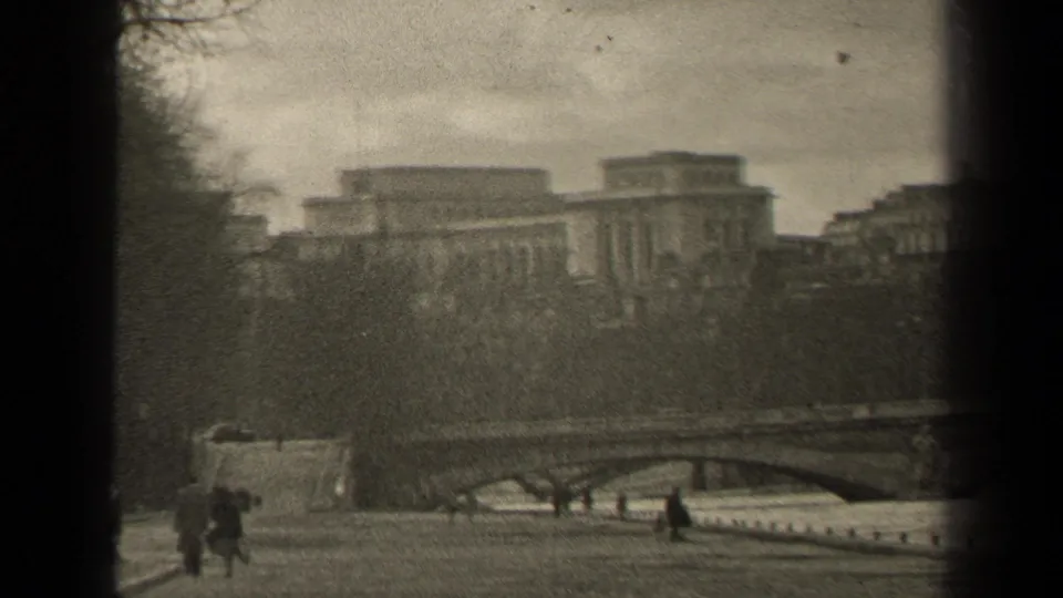 1947 Paris home-movie view across a riverside walkway toward pedestrians, bare trees, a bridge, and large civic-looking buildings.
