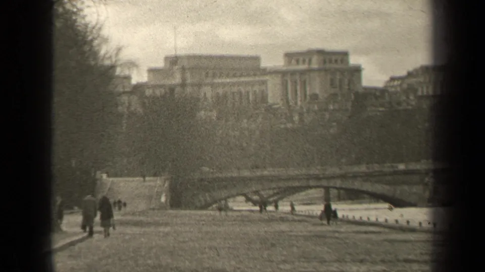 1947 Paris frame showing distant pedestrians on a riverside path, with a bridge, bare trees, and large buildings across the scene.