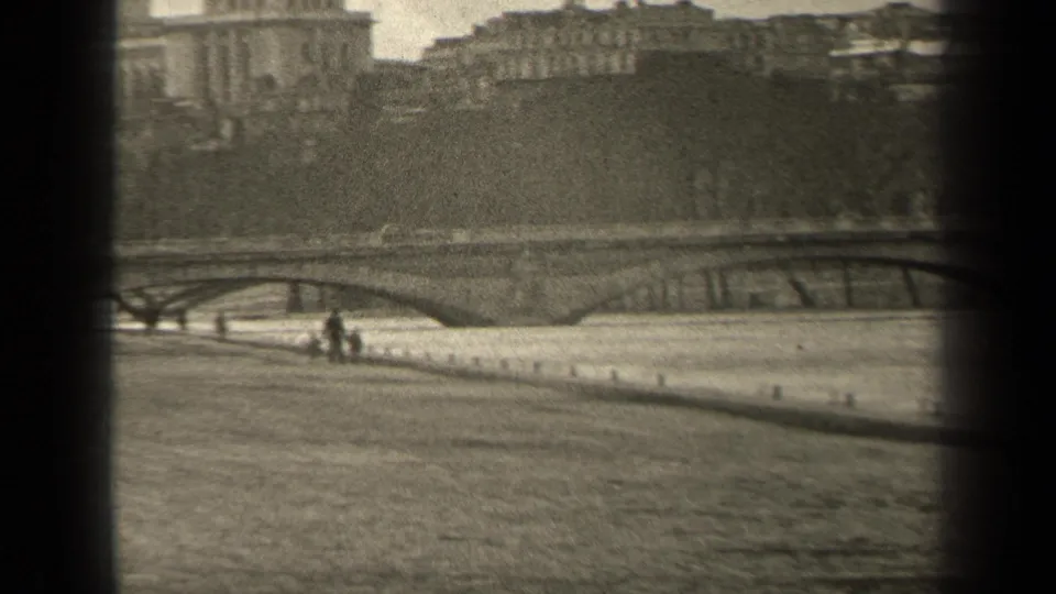 1947 Paris home-movie frame showing an arched stone bridge, riverside wall, and small pedestrians along the embankment.