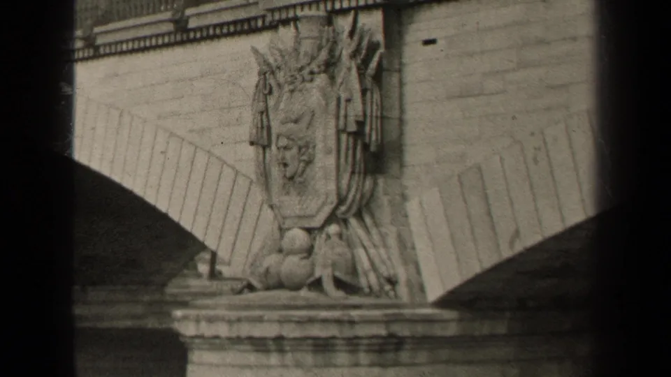 1947 Paris frame showing a sculpted bridge detail beneath a bare tree line, low fence, roadway, and distant urban rooflines.