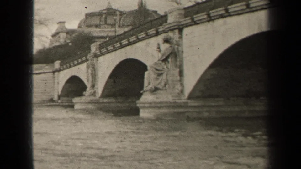 1947 Paris home-movie frame showing a stone arch bridge, river water, and a seated classical statue near the parapet.