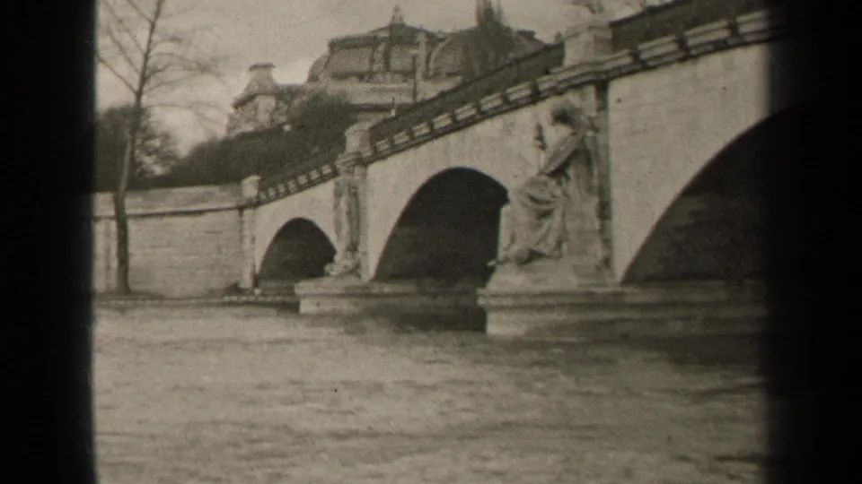 1947 Paris 8mm frame with bridge arches, a sculpted figure on a pedestal, bare trees, and a domed building beyond.