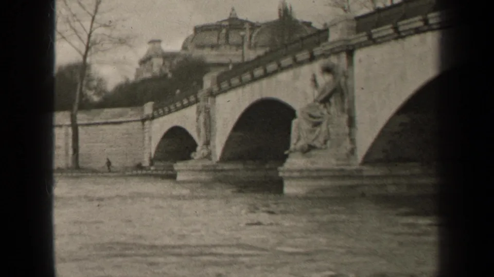 1947 Paris frame of a stone bridge over moving water, with a seated statue, bare trees, and historic buildings beyond.