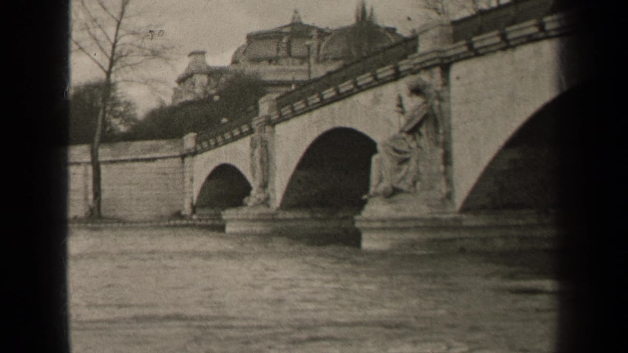 1947 Paris home-movie frame showing a stone arched bridge over water, with a seated statue on a pier and buildings behind.