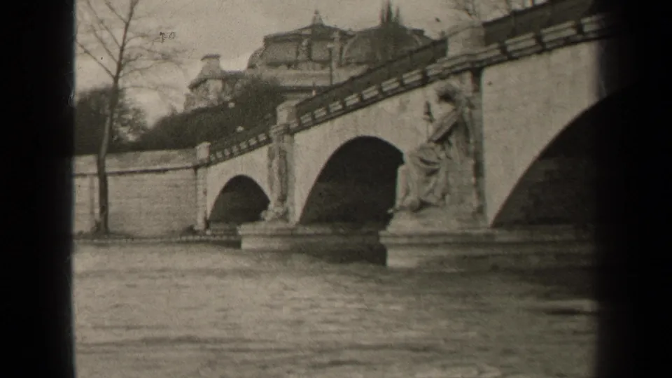 1947 Paris home-movie frame showing a stone arched bridge over water, with a seated statue on a pier and buildings behind.