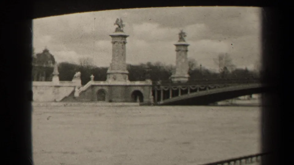 1947 Paris home-movie view across the river toward a bridge approach with tall sculptural pillars under an overcast sky.